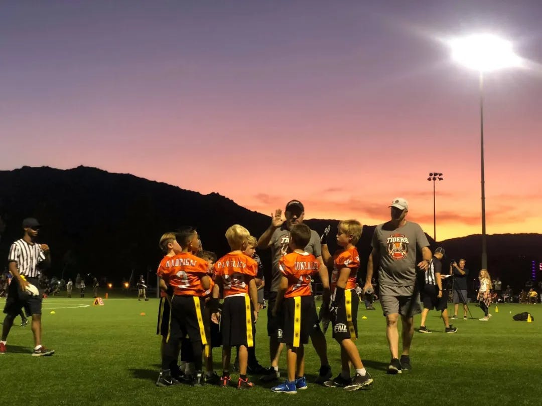 Image of sunset over a flag football field as the coach gives a high five to a bunch of kids