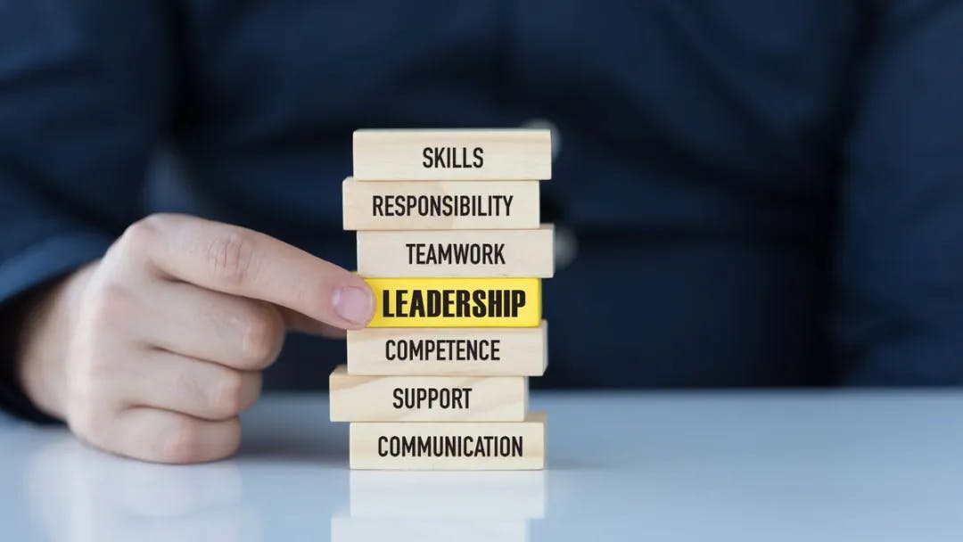 A hand holding a stack of wooden blocks, pointing to one that reads "leadership"