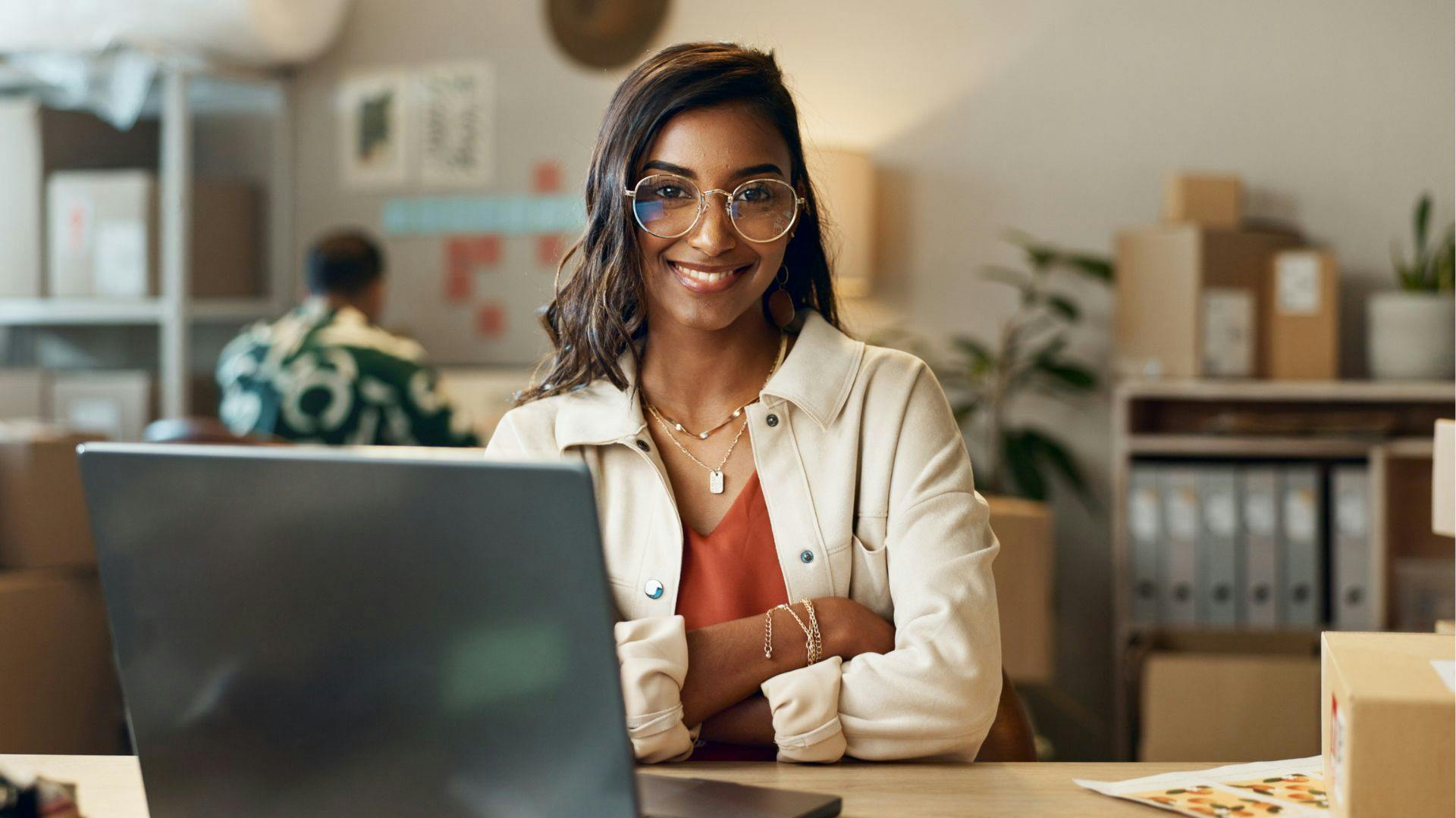 A woman wearing glasses is seated at a desk, working on a laptop computer.