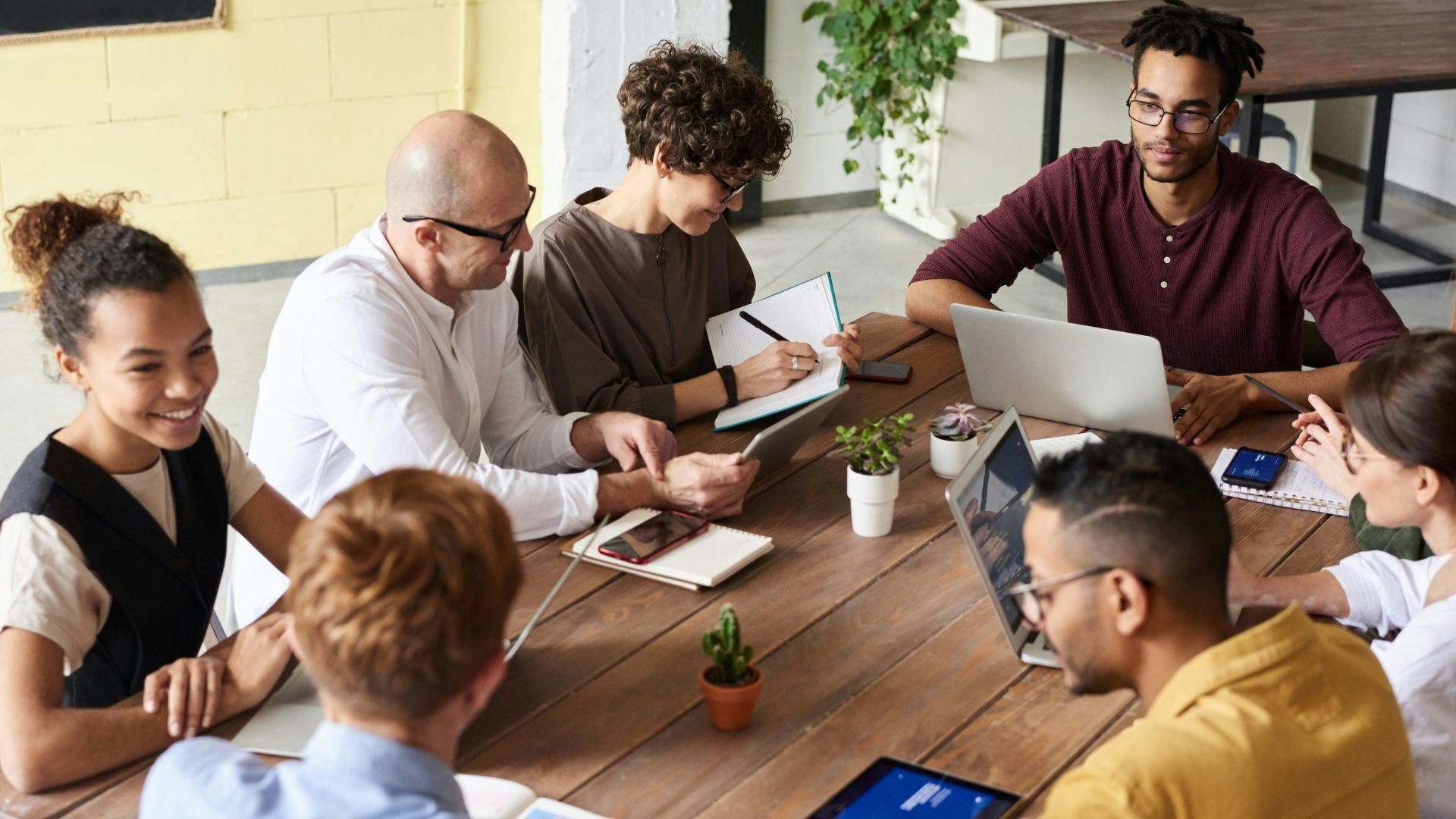 A diverse group of individuals sitting around a table with laptops, engaged in a meeting or collaboration session.