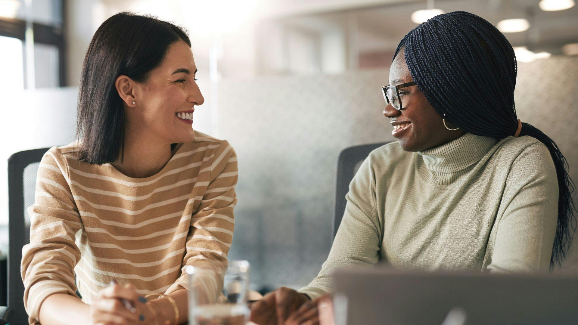 Two women conversing in an office, sharing ideas and collaborating in a professional setting.