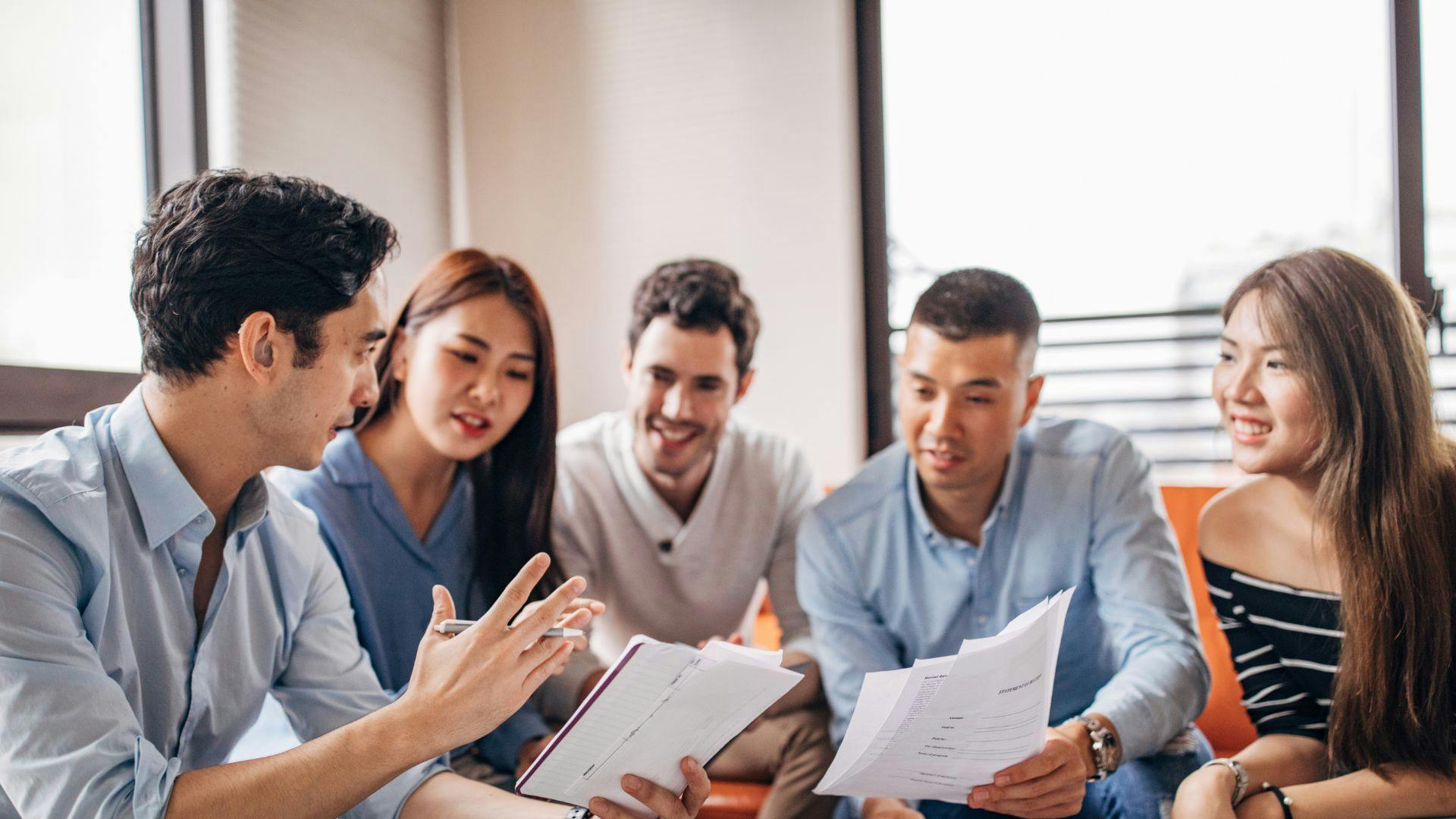 A team of young adults sits around a table, reviewing documents and exchanging ideas in a collaborative setting.