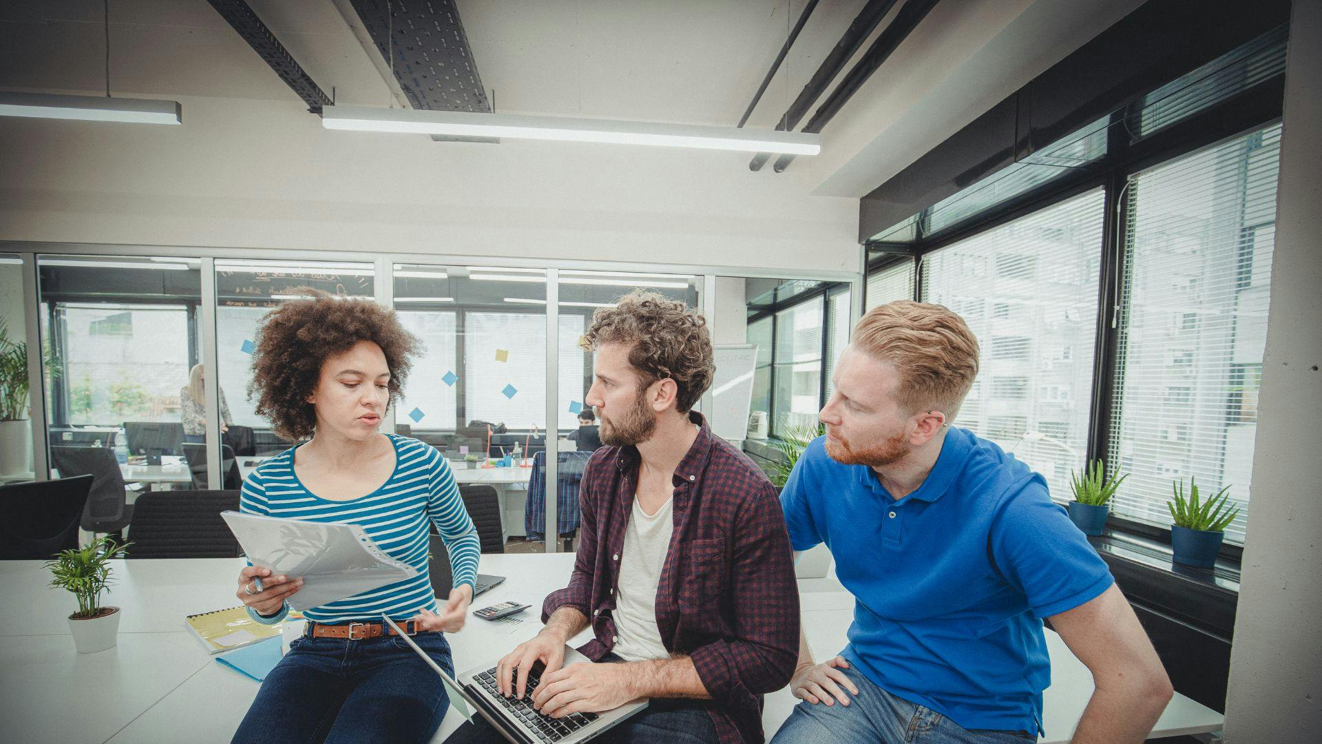 In an office setting, three people are gathered around a laptop, actively participating in a work-related conversation.