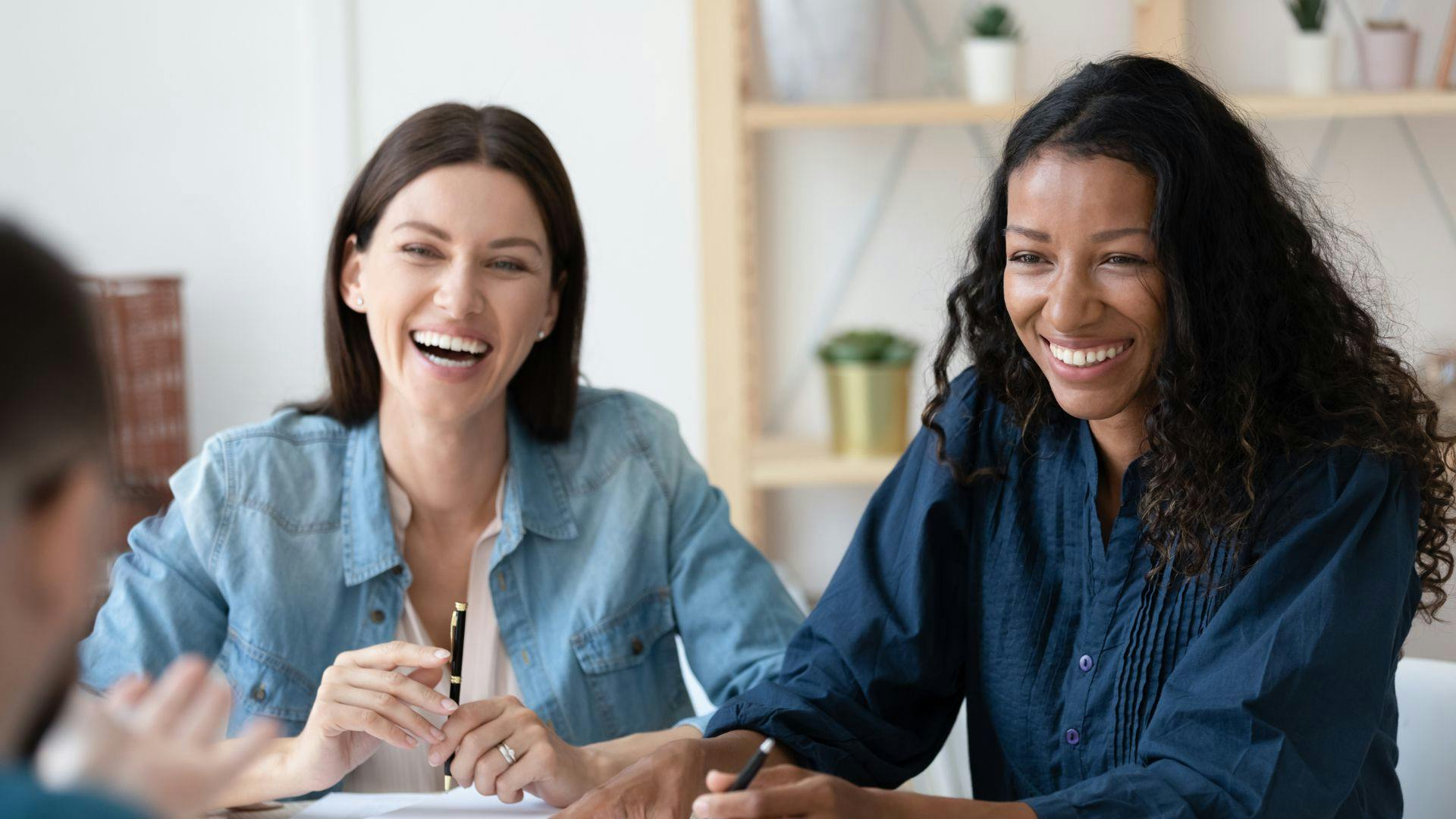 Two women laughing and smiling at a table during a friendly conversation.