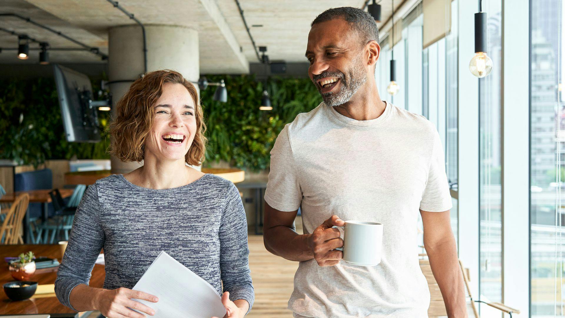 A cheerful man and woman enjoy a lighthearted conversation, both laughing and holding steaming cups of coffee.